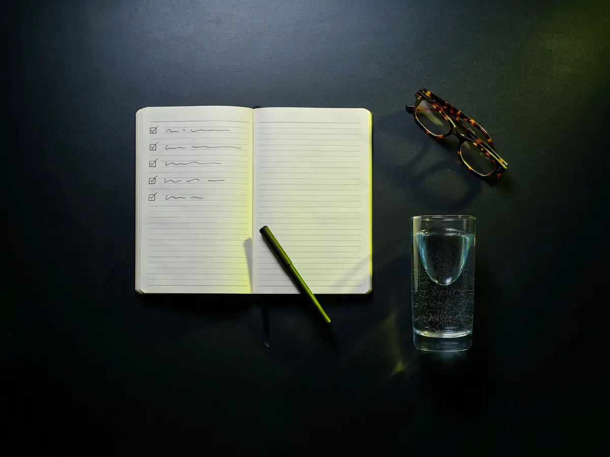 Ein aufgeschlagenes Notizbuch mit abgehakter Checkliste, ein Stift, eine Lesebrille und ein Glas Wasser